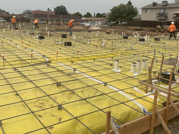 Construction site with workers, featuring a foundation with rebar grids and yellow waterproofing material, preparing for concrete pouring.
