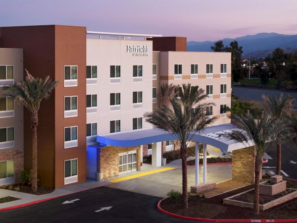 This image shows a modern four-story Fairfield Inn & Suites hotel with a front entrance, palm trees, and a parking area under a twilight sky.