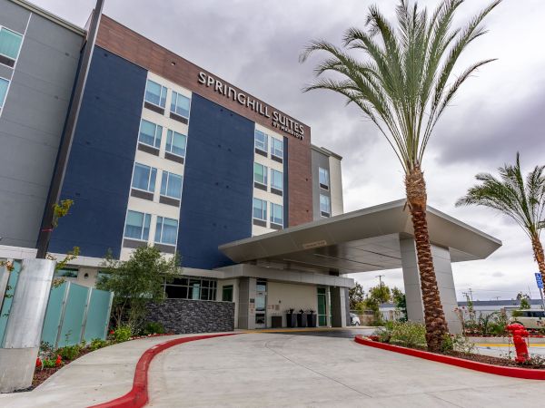 The image shows the exterior of a Springhill Suites hotel with a modern design, featuring palm trees and a driveway leading to the entrance.