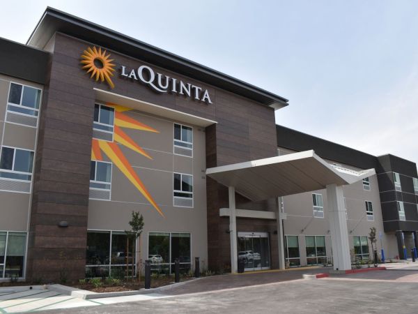 The image shows the exterior of a La Quinta hotel, featuring modern architecture with a prominent logo and a sundial design on the building.