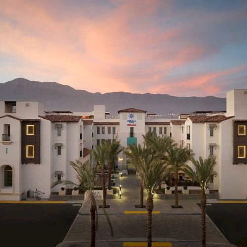 A building with Mediterranean architecture, surrounded by palm trees, set against mountains and a colorful sunset sky.