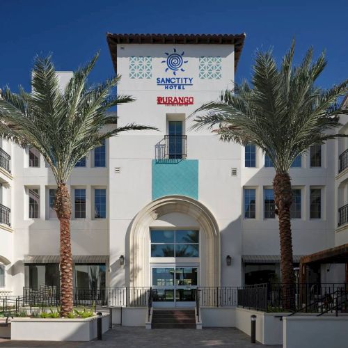 The image shows the front view of a hotel with palm trees, white walls, and the name "Sanctity Hotels" displayed.