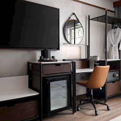 The image shows a modern hotel room corner with a TV, desk, minibar, chair, mirror, and robe by a window with curtains.