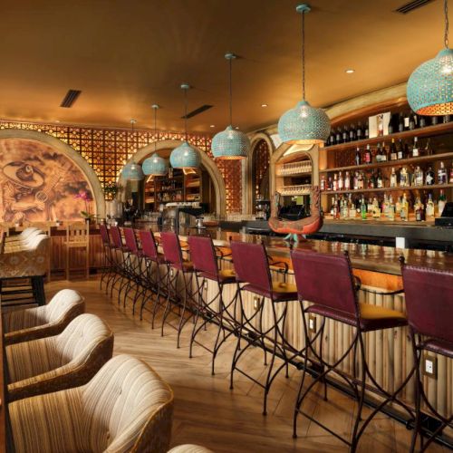 The image shows a cozy bar and restaurant with a mural, colorful lamps, high-backed barstools, and a variety of bottles and glasses on shelves.