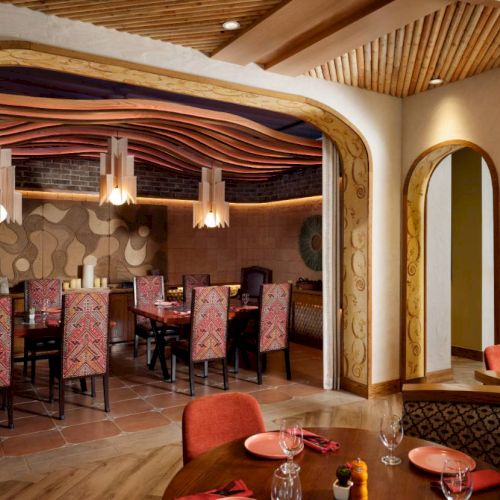 A warmly lit restaurant with colorful decor, patterned chairs, arched doorways, and a wooden ceiling. Tables are set with napkins and glasses.