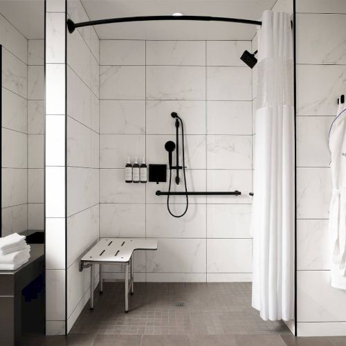 A modern, accessible bathroom with a tiled shower, a bench, wall-mounted bottles, a shower curtain, and a hanging robe.