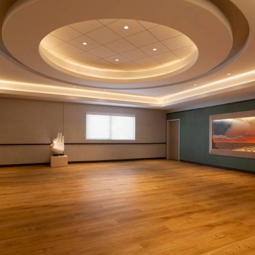 A serene room with a wooden floor, ceiling lights, a Buddha statue, a hand sculpture, and a sunset painting on the wall.
