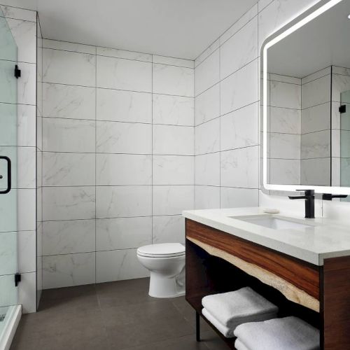 The image shows a modern bathroom with a glass shower, illuminated mirror, sink with storage, and a toilet against tiled walls.