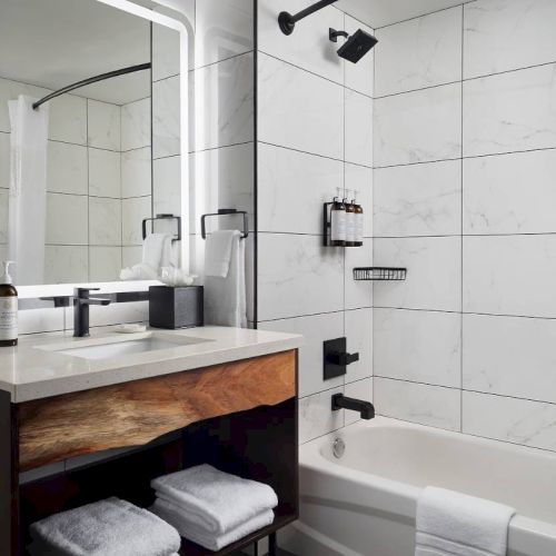 A modern bathroom with a lighted mirror, sink, wooden vanity, towels, shower/tub combo, and sleek black fixtures.