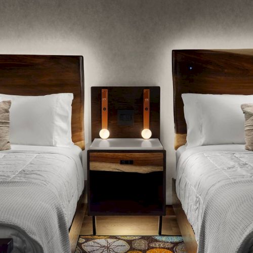Two beds with white linens and wood headboards, separated by a nightstand with lamps and a power outlet.