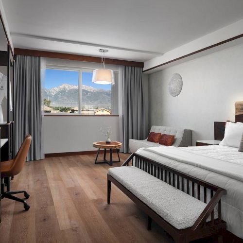 A hotel room with a bed, desk, chair, and a large window with mountain views. Modern decor includes a sofa, table, and wall art.