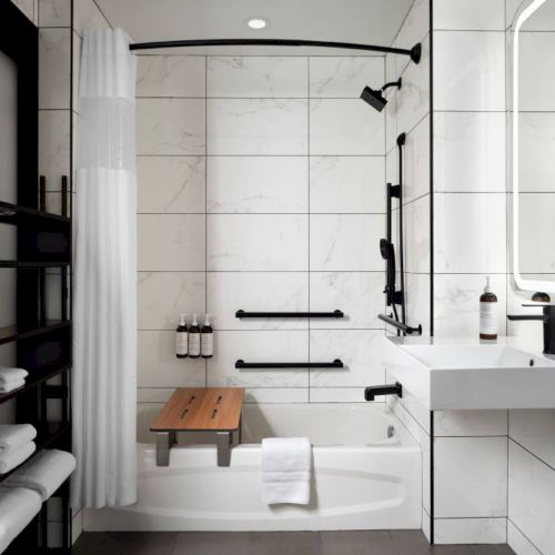 The image shows a modern bathroom with a bathtub, white tiles, a black shower fixture, shelves with towels, and a large mirror.
