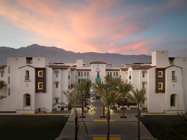 The image shows a large white building with palm trees in front, against a backdrop of mountains and a colorful sunset sky.