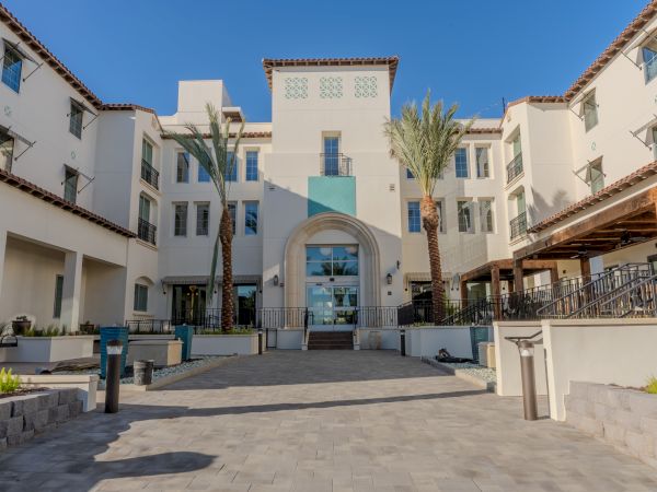 A modern Mediterranean-style building with palm trees, arched entryway, and a spacious courtyard under a clear blue sky.