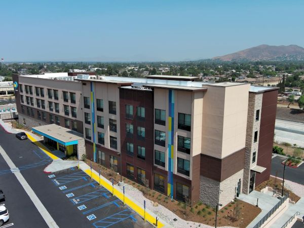 The image shows a modern, multi-story building, likely a hotel or an apartment complex, with parking spaces and a cityscape in the background.
