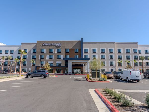 The image shows a modern Hampton Inn & Suites hotel with a parking lot and a few vehicles in front.
