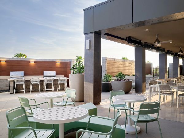The image shows an outdoor patio with tables, chairs, and grills surrounded by modern decor and plants under a clear sky.