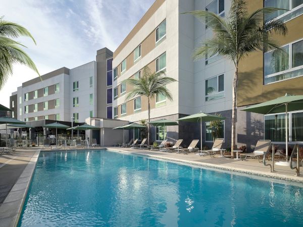 A modern hotel with an outdoor pool, surrounded by sun loungers and palm trees, against a clear sky backdrop.
