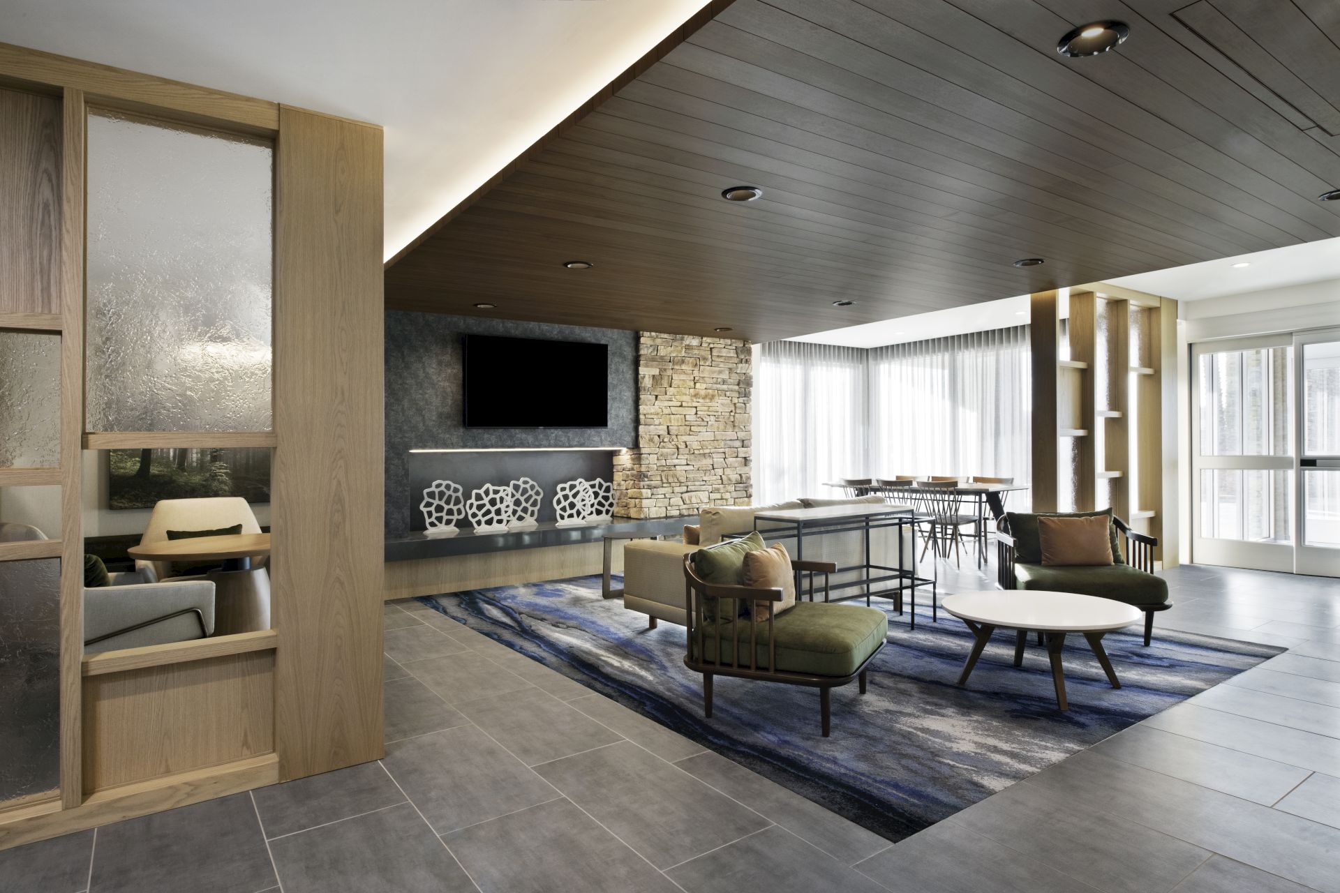 A modern lounge area with stylish furniture, a large rug, and a wall-mounted TV. The room has ample natural light from large windows.