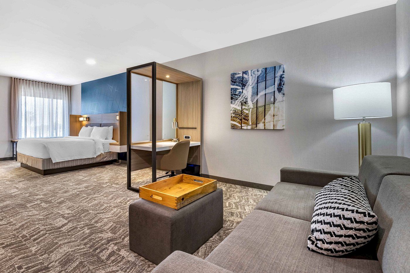 A modern hotel suite featuring a bed, desk, sofa, and divider. The room is well-lit with a window, lamp, and has abstract art on the wall.