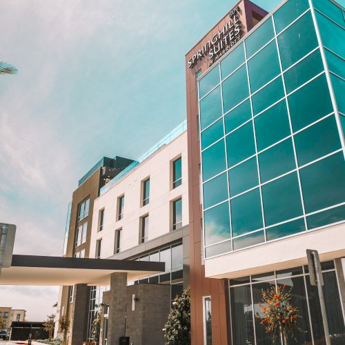 A sunny city street with modern teal-blue building, palm trees, and a few pedestrians, giving a bright, tropical urban vibe.