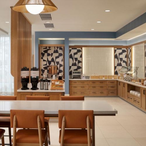 A bright, modern breakfast area with a long counter, coffee setup, and seating; warm wood tones and patterned wall panels.