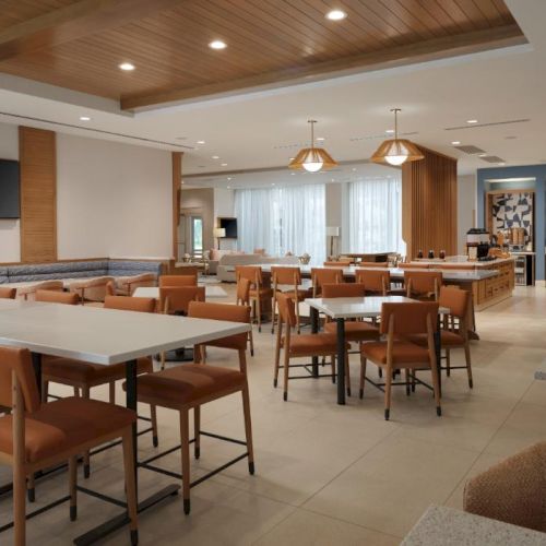 A modern, well-lit dining/lounge area with wooden tables and chairs, a large screen, and cozy seating along the walls, in a hotel or cafe.