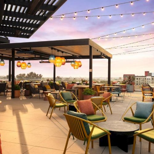 Rooftop bar with string lights, colorful chairs, and marble tables overlooking a sunset skyline, perfect for socializing and cocktails.