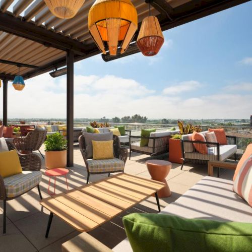 A bright rooftop lounge with wicker seating, colorful cushions, and hanging lanterns overlooking a sunny cityscape, perfect for relaxing al fresco.