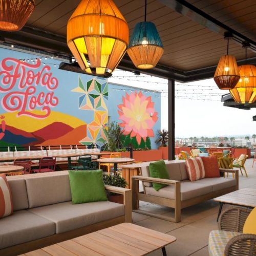 A bright rooftop lounge with colorful sofas, woven chairs, and vibrant throw pillows, a mural says &ldquo;Flower Love,&rdquo; and hanging lanterns glow above.