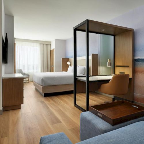 A stylish hotel room with a split seating area, a bed, a glass-walled workspace, wooden floors, and a beach mural, bright and modern.