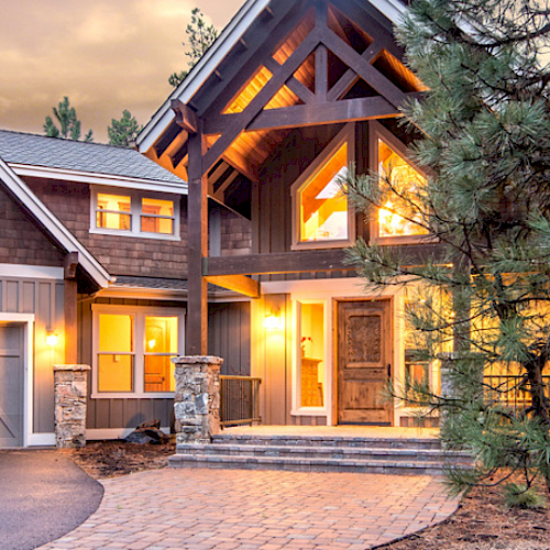 A large, modern home with glowing windows and a driveway, surrounded by trees in the evening.