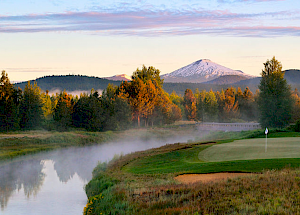 A beautiful golf course by a misty river surrounded by trees, with mountains in the background under a serene sky.