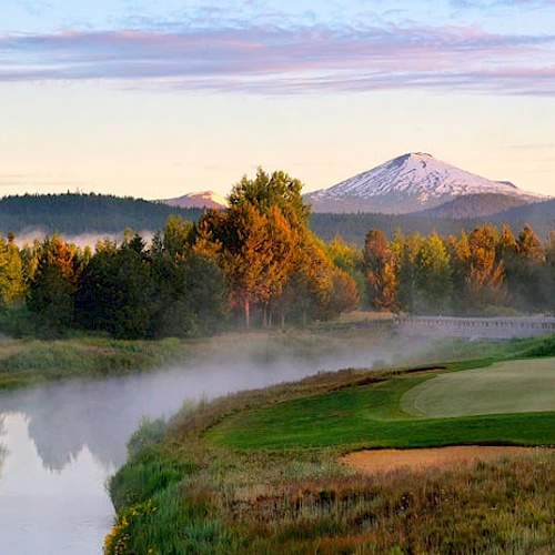 A beautiful golf course by a misty river surrounded by trees, with mountains in the background under a serene sky.