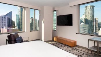 A modern hotel room with city views through large windows, featuring a bed, armchair, wall-mounted TV, wooden cabinet, and small table with a chair.