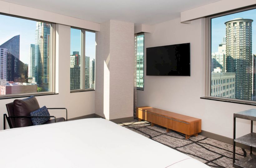 A modern hotel room with city views through large windows, featuring a bed, armchair, wall-mounted TV, wooden cabinet, and small table with a chair.