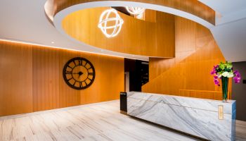 Modern hotel lobby with a marble reception desk, wooden spiral staircase, large wall clock, and decorative light fixtures and flowers.