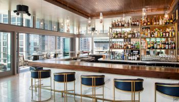 A modern bar with a long counter, stylish barstools, a backdrop of shelves filled with various bottles, and large windows showcasing a cityscape.
