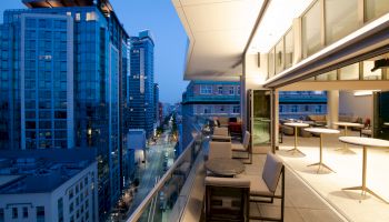 A rooftop terrace at twilight, with modern seating, glass railings, and city buildings in view. Indoor area opens to the terrace with lights on.