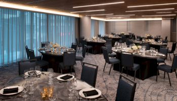 A spacious banquet room set with round tables covered in white flowers, black napkins, and silverware, illuminated by modern overhead lighting.