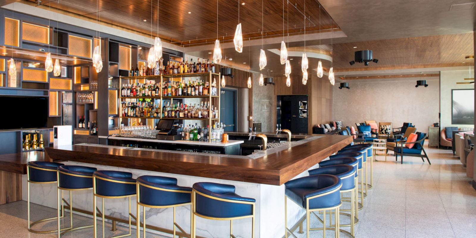 The image depicts a modern bar with blue barstools, hanging lights, and a well-stocked shelf of bottles behind the counter, set within a spacious lounge area.