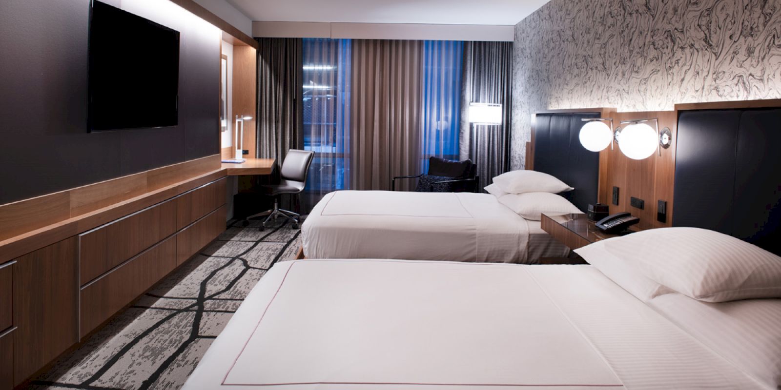 A modern hotel room with two beds, a wall-mounted TV, a work desk, two chairs, and stylish decor.