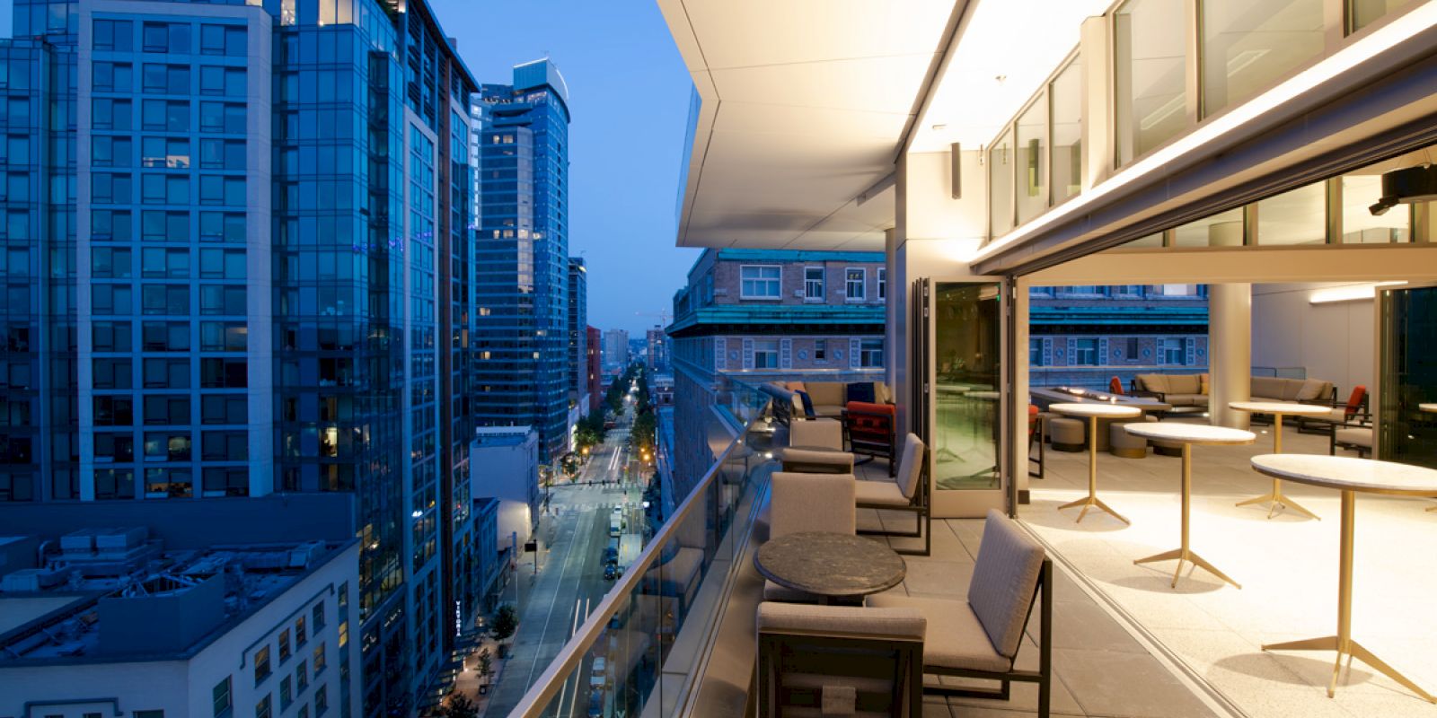 The image shows a modern urban balcony at dusk, with outdoor seating and tables overlooking a city street lined with tall buildings.