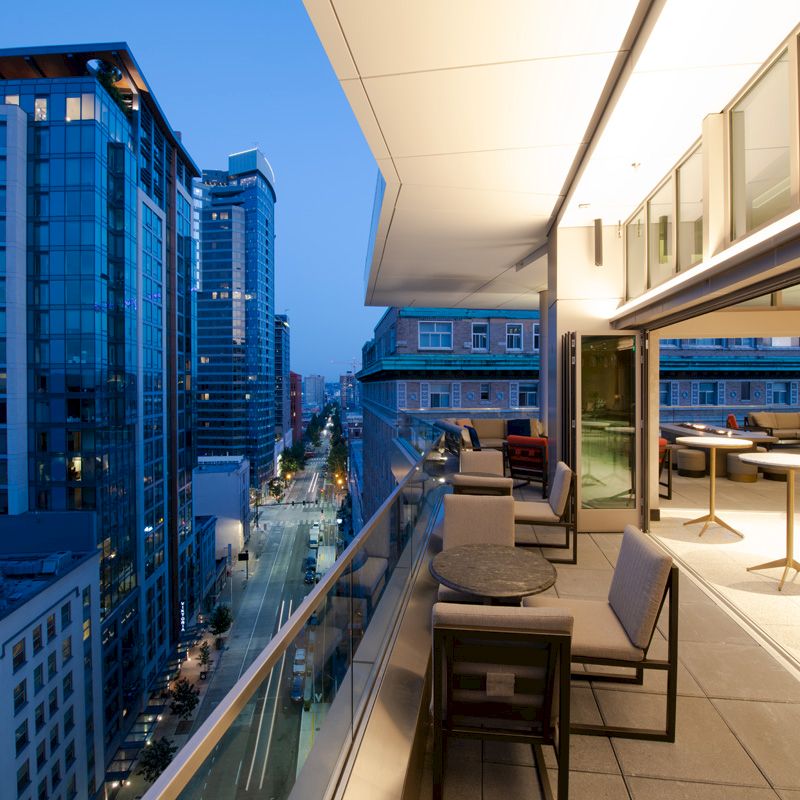 A modern balcony with seating and tables, overlooking a cityscape featuring tall buildings and a busy street below, captured during twilight.