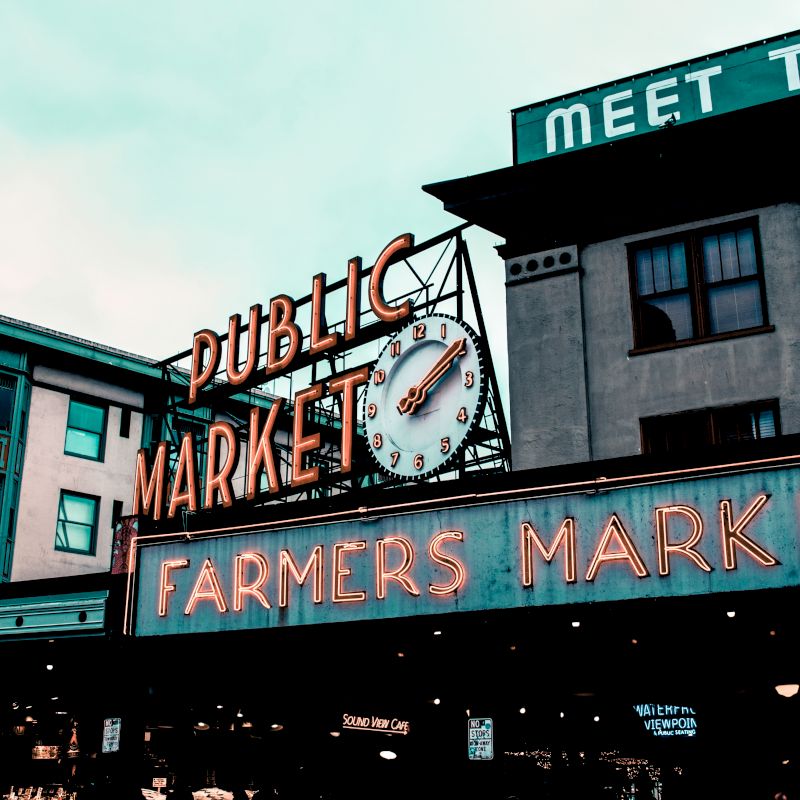 A public market with neon signs reading "Public Market" and "Farmers Market," along with a clock. Surrounding buildings add to the urban atmosphere.