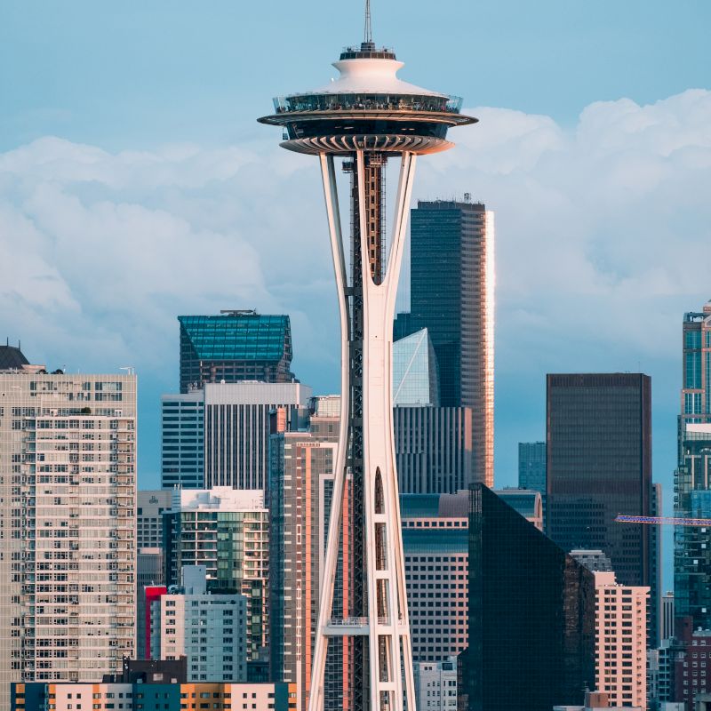 The image shows the Seattle Space Needle against a backdrop of the city's skyscrapers and cloudy sky. It captures the urban skyline in daylight.