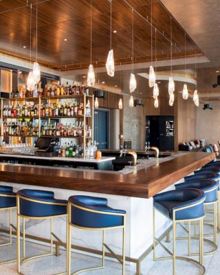 The image showcases a modern bar with a wooden countertop, stylish pendant lighting, blue barstools, and a well-stocked liquor shelf in a spacious setting.