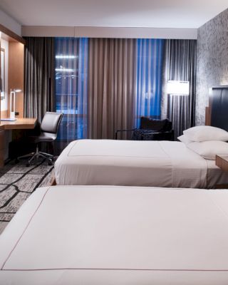 The image shows a modern hotel room with two neatly made beds, a desk with a chair, a wall-mounted TV, and contemporary decor.