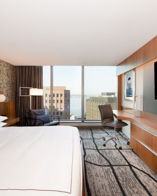 A modern hotel room with a large bed, a desk with a chair, a flat-screen TV, and a view of buildings and water through a large window.