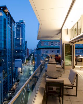 This image shows a modern outdoor balcony area with seating, overlooking a cityscape with tall buildings and a street below during the evening.